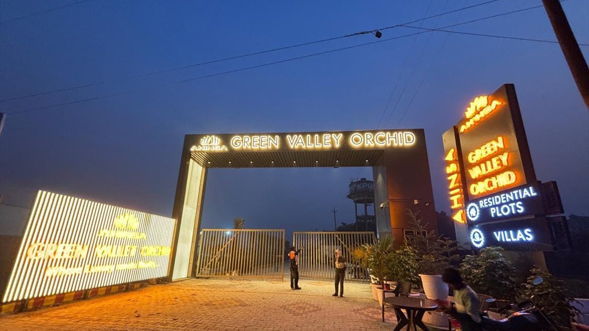 The lit entrance gate of Green Valley Orchid at dusk, beside a hoarding advertising residential plots and villas.