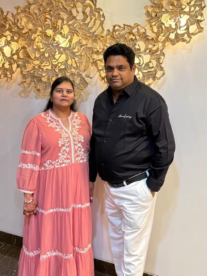 Rohit Jain and his wife standing in front of a wall of gilded floral sculptures; she wears a pink embroidered dress, he a black shirt and white trousers.