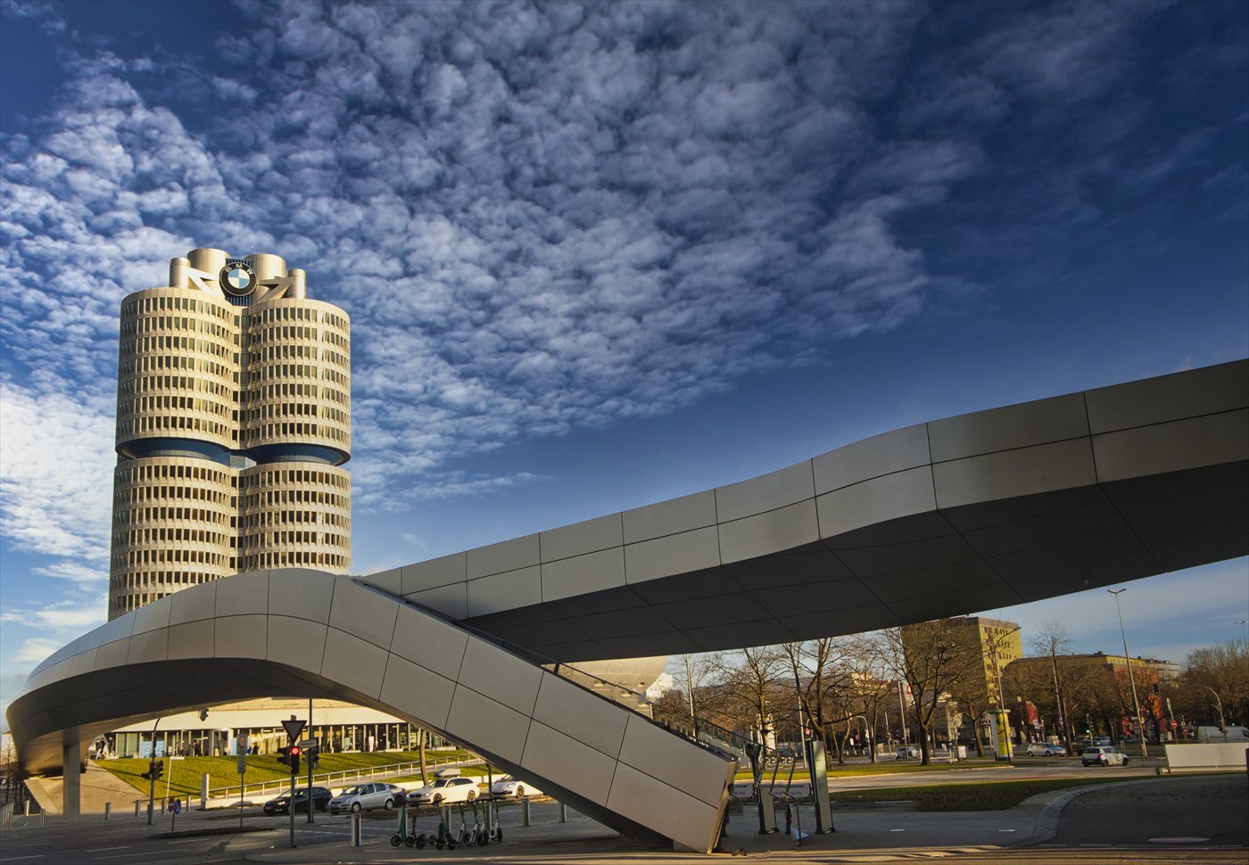 BMW’s cylindrical headquarters tower in Munich beside the sweeping steel-and-glass roof of BMW Welt, under a blue sky with clouds.
