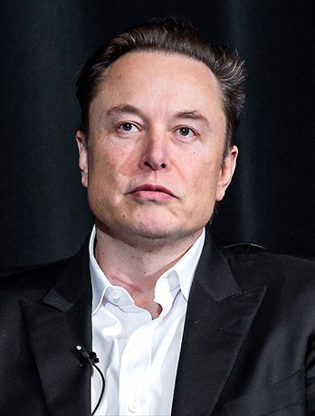 Elon Musk in a dark jacket and open-collared white shirt, photographed at a speaking event.