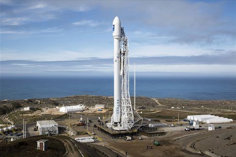 A white SpaceX Falcon 9 rocket standing vertical on a coastal launch pad with the Pacific behind it.