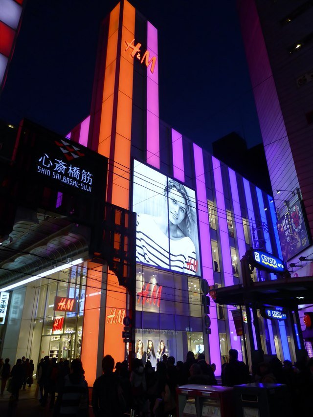 An H&M store in the Shinsaibashi shopping district of Osaka at night, its tall front lit in orange, purple and blue.