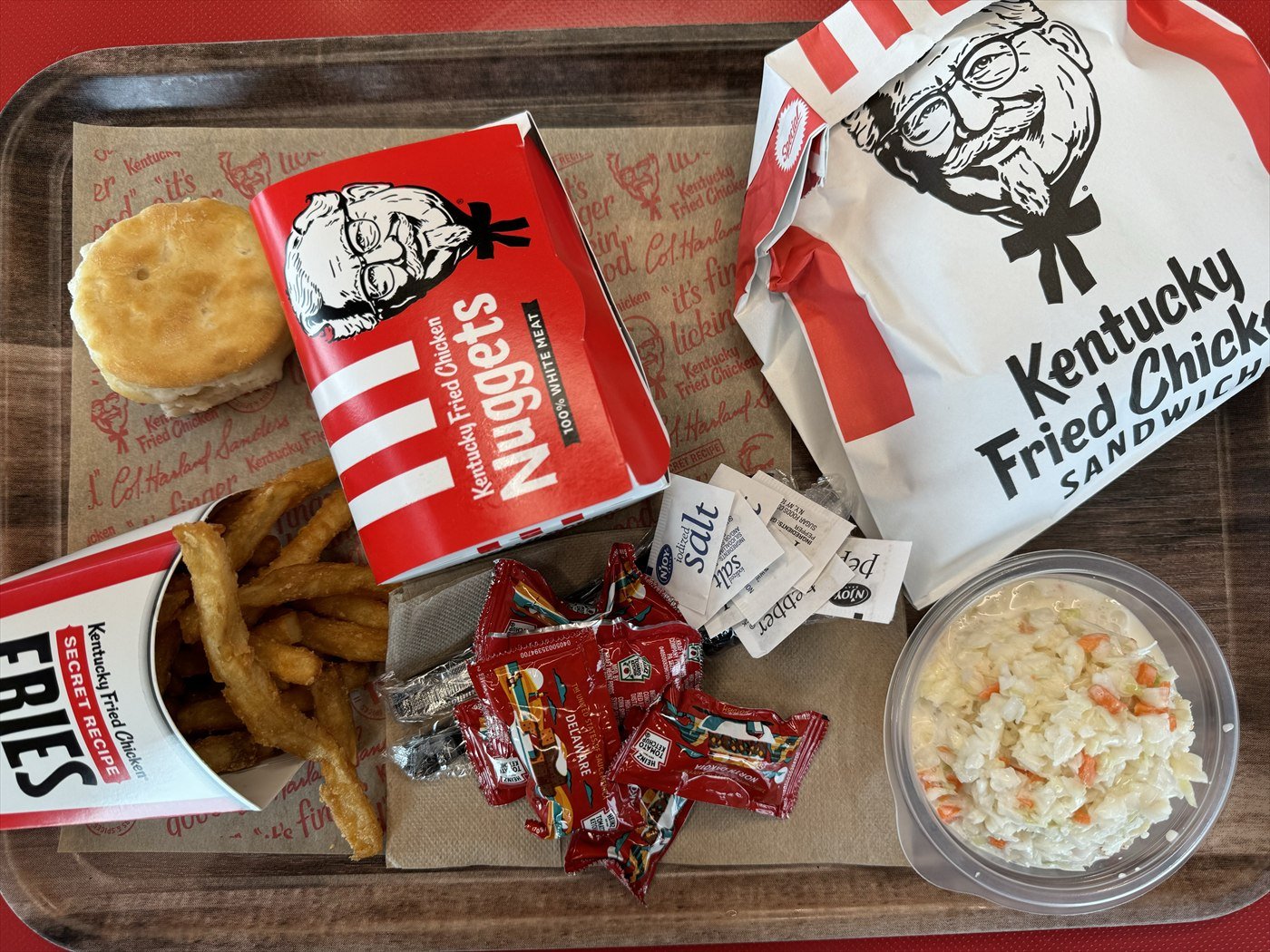 A KFC meal on a tray: a chicken sandwich in a Kentucky Fried Chicken bag, nuggets, fries, a biscuit, coleslaw and sauce packets.