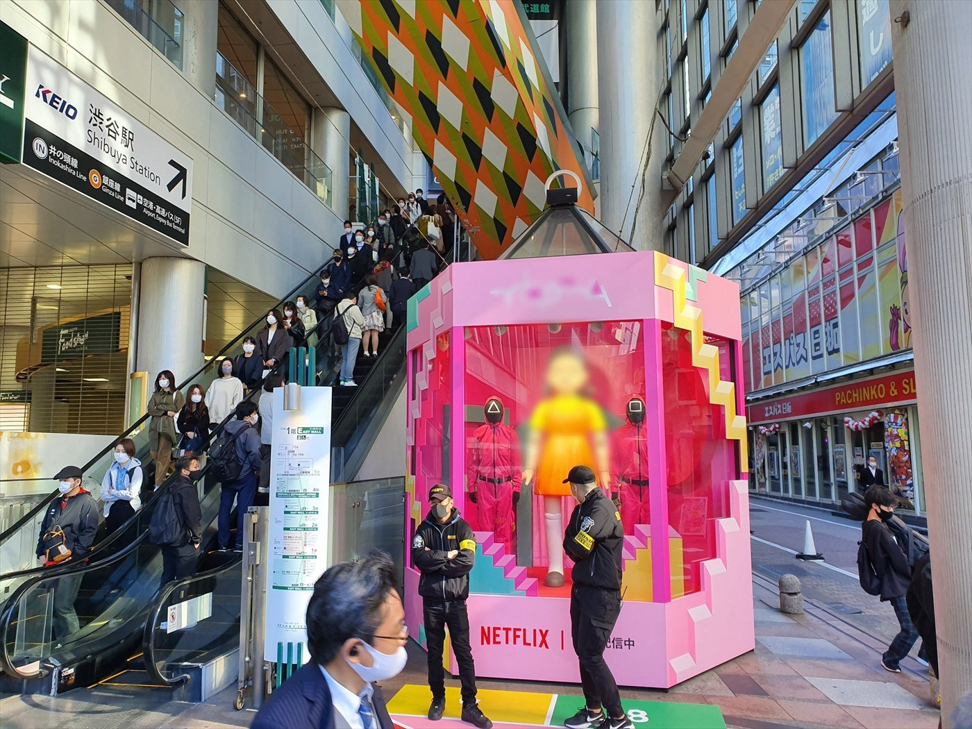 A pink Squid Game display booth outside Shibuya Station in Tokyo, with two masked guard figures in pink jumpsuits, a blurred doll figure and the Netflix logo, as commuters ride an escalator beside it.