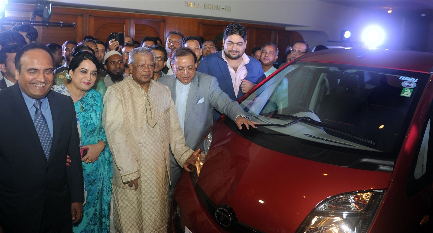 A red Tata Nano on display indoors, surrounded by guests at a launch event, the Tata badge visible on its bonnet.