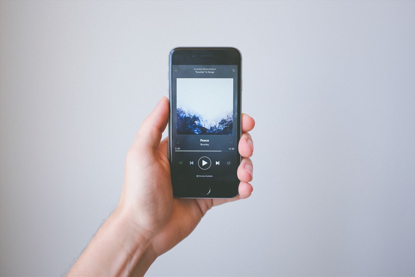 A hand holding up a smartphone showing a song playing in the Spotify app, against a pale grey background.