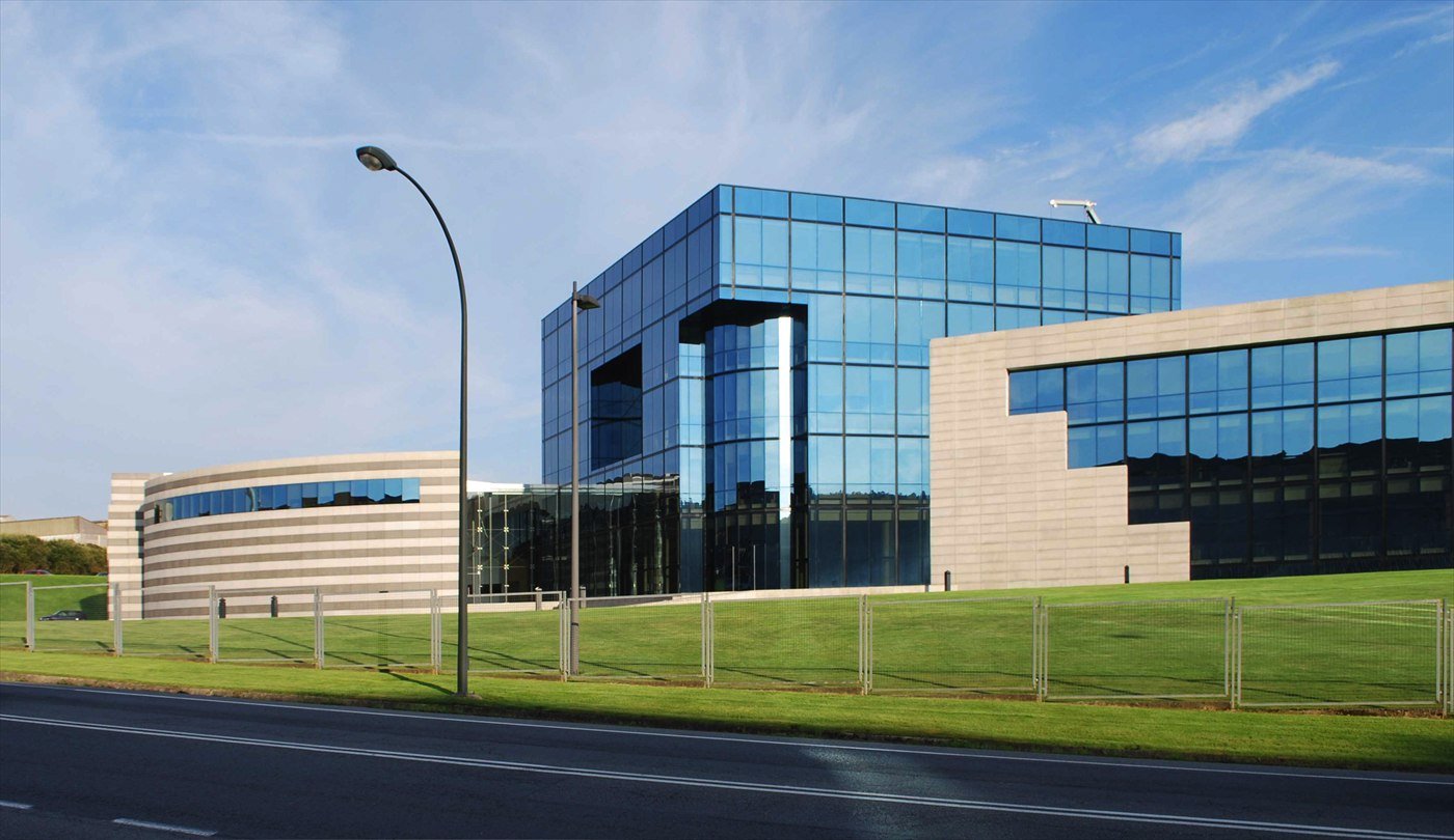 Inditex’s headquarters in Arteixo: a modern glass-and-stone office building beside a lawn and a road under a blue sky.